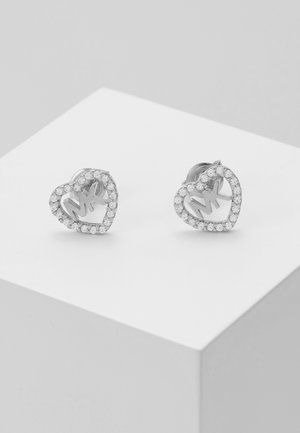 Boucles d’oreilles en argent en forme de cœur, ornées de cristaux clairs délimitant le contour du cœur et des initiales "MK" au centre, présentées sur une surface blanche.
