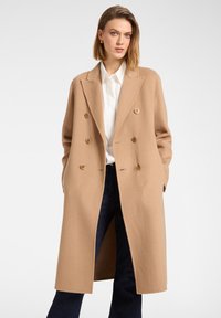 Mujer con un abrigo largo camel de doble botonadura, camisa blanca y pantalones oscuros, con las manos en los bolsillos del abrigo sobre un fondo claro y liso.