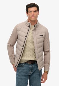 Veste rembourrée beige clair avec un design matelassé, fermeture à pression et col montant, portée sur un pull en maille torsadée crème et un jean bleu.