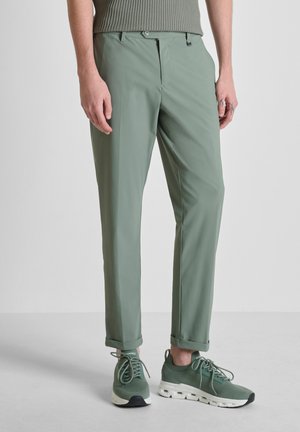 Uomo che indossa pantaloni su misura verde salvia con orli risvoltati e sneakers verdi abbinate, in piedi su un pavimento semplice.