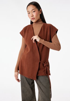 Giovane donna con lunghi capelli scuri indossa un gilet avvolgente senza maniche marrone sopra un dolcevita a coste abbinato e pantaloni grigi a gamba larga, in posa.