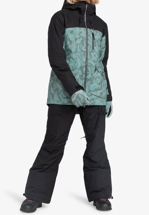 Persoon die een zwarte en tealbloemenprint winterjas draagt, bijpassende handschoenen, zwarte sneeuwbroek en zwarte sneeuwlaarzen.