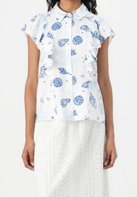 Donna che indossa una blouse bianca a maniche corte con stampe di conchiglie blu e rouches, abbinata a una gonna bianca in pizzo texturizzato.
