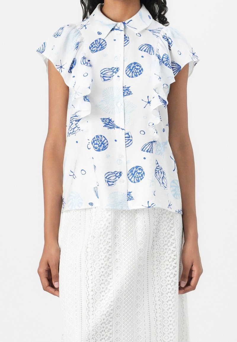 Donna che indossa una blouse bianca a maniche corte con stampe di conchiglie blu e rouches, abbinata a una gonna bianca in pizzo texturizzato.