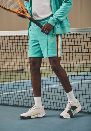 Homme en tenue de sport bleu-vert tenant une raquette de tennis orange, debout sur un court de tennis bleu près d'un filet noir, portant des baskets blanches avec des touches jaune-vert.