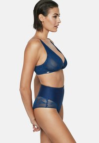 Femme aux cheveux courts et mouillés portant un bralette en dentelle bleu marine et des culottes en dentelle taille haute, se tenant de profil sur fond blanc.