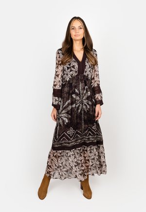 Woman stands wearing a long-sleeve, V-neck black and white patterned maxi dress with brown heeled boots against a plain white background.
