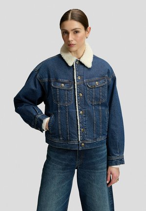Frau mit braunen Haaren, die eine blaue Jeansjacke mit weißem Sherpa-Kragen und blaue Jeans trägt, steht vor einem schlichten hellen Hintergrund.