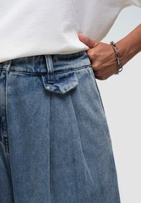 Pantalones vaqueros en azul claro con un frontón plisado, que cuentan con un cierre de botón y cremallera, y un pequeño bolsillo de parche cerca de la cintura.