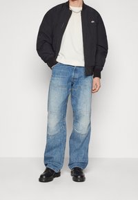 Schwarze Bomberjacke, weißes T-Shirt, hellblaue Jeans mit Patchwork, lockere Passform und schwarze Schnürstiefel. Silberne Halskette sichtbar.