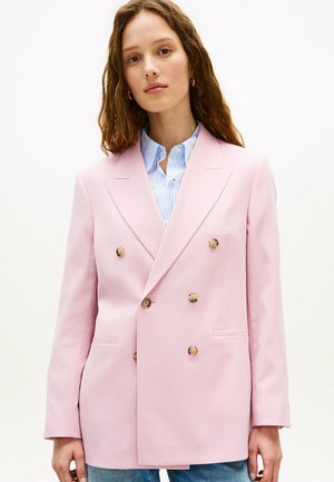 Kvinde med langt brunt hår iført en lyserød dobbeltradet blazer over en blå og hvidstribet skjorte og blå jeans, stående mod en hvid baggrund.