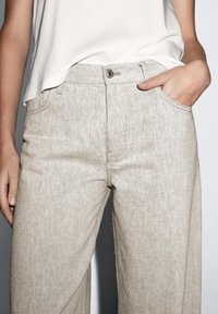 Helle beige weit geschnittene Hose aus strukturiertem Stoff, mit einer Vordertasche, einem einfachen Knopfverschluss und klaren Nahtdetails.