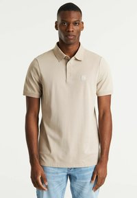 Polo beige in tessuto testurizzato, maniche corte, colletto a punta e un piccolo logo sul petto sinistro, abbinato a jeans azzurri chiari.