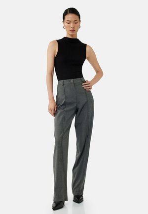 Un haut sans manches noir associé à un pantalon gris ajusté, avec un bouton à la taille et de subtiles plis, porté avec des chaussures noires élégantes.