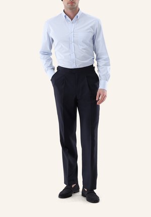 Mann trägt hellblaues Hemd mit langen Ärmeln, dunkelmaßgeschneiderte Hose mit Falten und schwarze Slipper, steht mit einer Hand in der Tasche.