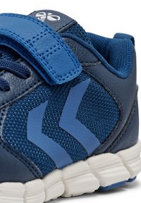 Zapato atlético azul con una parte superior de malla texturizada y acentos de cuero, correa ajustable con gancho y bucle, y una suela de goma blanca con ranuras de tracción.
