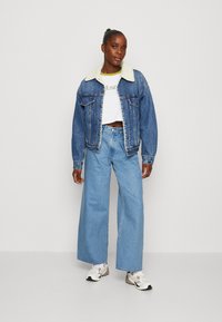 Jeans jack met een schapenvachtkraag, gecombineerd met een cropped witte T-shirt en wijde lichtblauwe jeans. Witte en beige sneakers maken de look compleet.