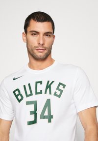 Hvid t-shirt med kortærmer, der har "BUCKS" og "34" trykt i grønne og gyldne bogstaver. Nike-logo i grøn på venstre skulder. Materiale: bomuld.