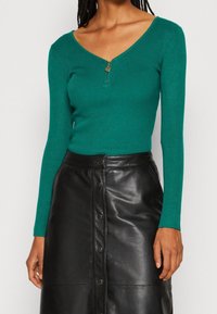 Haut vert côtelé à manches longues avec un col en V et un détail de fermeture éclair à l'avant, associé à une jupe en cuir noire avec des boutons.