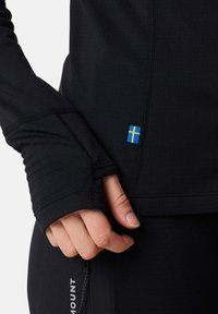 Musta pitkähihaine urheilu paita, jossa on peukalonreiät, varustettuna pienellä sinisen ja keltaisen sävyisellä Ruotsin lipun tarralla. Silkkinen pinta ja tasaiset saumat.
