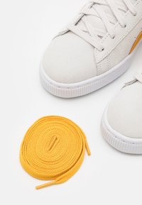 Detailní záběr na bílé tenisky s navinutou žlutou tkaničkou položenou vedle nich na bílé ploše.