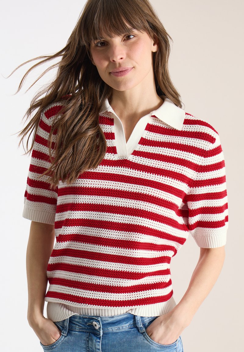 Jeune femme aux longs cheveux bruns portant un polo tricoté à manches courtes à rayures rouges et blanches et un jean bleu, les mains dans les poches.