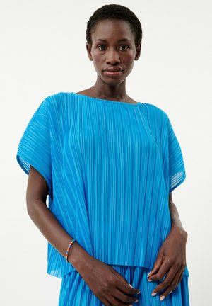 Faltige blaue Bluse mit weiten Ärmeln, aus leichtem Material gefertigt. Das Modell steht mit vor der Brust gefalteten Händen und präsentiert den strukturierten Stoff.