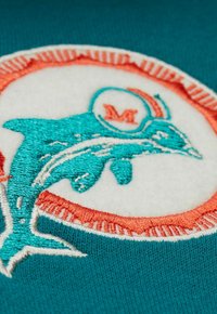 Türkiser Stoff mit einem gestickten Delfin-Motiv in Türkis und Weiß, umrandet von einem orangefarbenen Rand mit kleinen Akzenten.