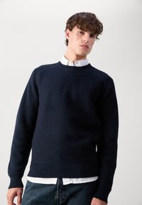 Felpa blu navy con maniche e orlo a costine, caratterizzata da una finitura testurizzata e un design a V, indossata sopra una camicia bianca con colletto.