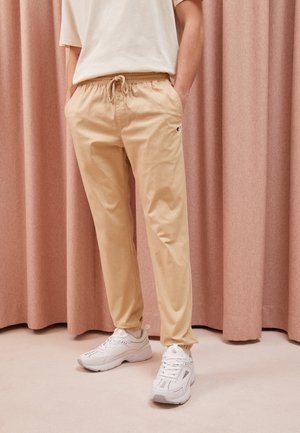 Pantalons jogger en coton beige avec une taille élastique et un cordon de serrage. Présente un petit logo sur la cuisse gauche. Portés avec des baskets blanches.