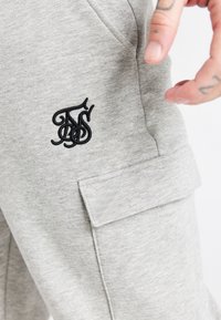 Szare dresy wykonane z mieszanki bawełny, z czarnym haftowanym logo na udzie oraz boczną kieszenią z klapką.