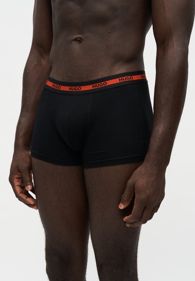 Buste masculin portant un boxer noir avec une ceinture rouge affichant le nom de la marque HUGO en lettres noires.