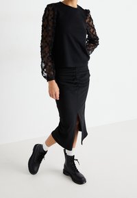 Svart topp med genomskinliga ärmar broderade med blommönster, kombinerad med en svart midjekjol med rynkdetalj och svarta snörstövlar med vita strumpor.