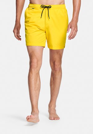 Man met felgele zwemshorts met een zwarte trekkoord, staat blootsvoets op een effen witte achtergrond.