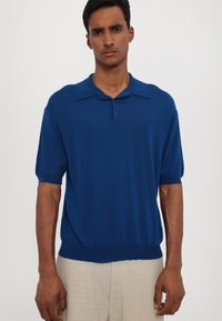 Niebieska dzianinowa koszulka polo z kołnierzykiem, krótkimi rękawami i zapięciem na dwa guziki. Noszona z jasnymi, teksturowanymi beżowymi spodniami.