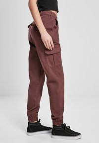 Burgundské cargo kalhoty z látky s elastickými manžetami, bočními kapsami a volným střihem. Nosené s černými vysokými teniskami.