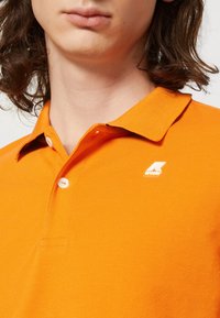 Personne portant un polo orange avec des boutons blancs et un petit logo K-Way blanc et orange sur la poitrine.
