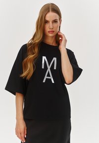 Czarna, oversize t-shirt z krótkim rękawem, z wyrazistym białym graficznym napisem "MA" na przodzie. Gładki materiał bawełniany.