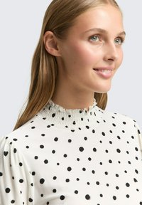 Weißes Oberteil mit schwarzen Pünktchen, versehen mit einem gesmokten hohen Kragen und Puffärmeln, aus einem glatten Stoff gefertigt.