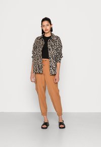 Camisa em estampa de leopardo, blusa preta e calças cargo castanhas com punhos elásticos. Sandálias pretas; modelo de pé sobre um fundo neutro.