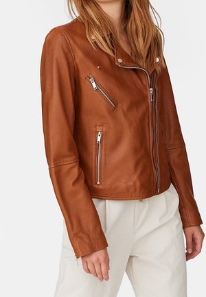 Woman wearing a fitted brown leather jacket with zipped pockets and silver hardware, paired with high-waisted beige pants.