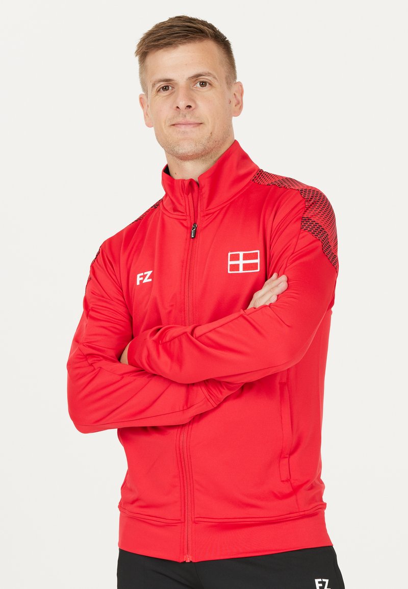 Röd idrottsjacka med hög krage, hel dragkedja, logotyp på bröstet och strukturerat mönster på axlarna. Tillverkad av ett mjukt, lätt material.
