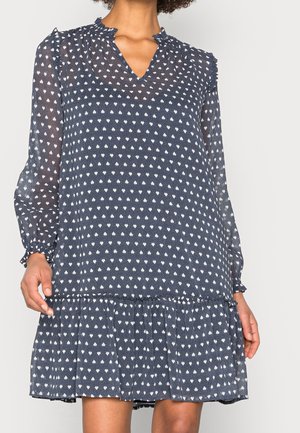 Vestido azul marino de manga larga con patrón de diamantes blancos, cuello y dobladillo con volantes, llevado por una persona con tono de piel medio oscuro.