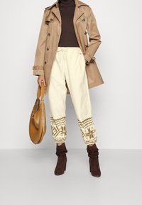 Beige trenchcoat över en brun turtleneck, parat med krämfärgade fleecebyxor med ett geometriskt mönster vid fållarna och bruna klackstövlar.