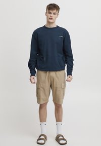 Mornarica pulover z okroglim izrezom in sprednjim žepom, v kombinaciji s bež cargo kratkimi hlačami s stranskimi žepi, belimi nogavicami in rjavimi sandali.