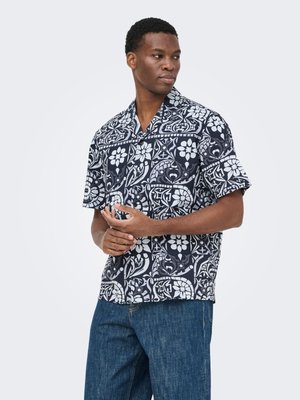 Homme portant une chemise à manches courtes boutonnée avec un imprimé floral et géométrique noir et blanc, associée à un jean bleu, debout devant un fond uni.