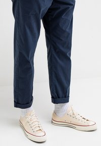 Navyblauwe getailleerde broek met opgerolde manchetten, gecombineerd met beige canvas sneakers en lichtgrijze geribbelde sokken met rode en blauwe accenten.