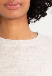 Gesicht und Hals einer Frau mit mittelbrauner Haut, die einen hellbeigen, weichen Strickpullover trägt, während ihre lockigen, dunklen Haare an der Seite sichtbar sind.