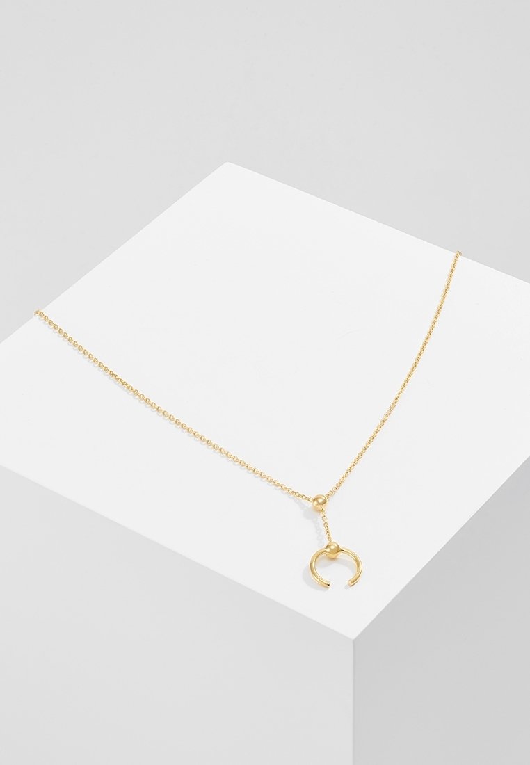 Collier délicat en chaîne d'or avec un petit pendentif circulaire ouvert et deux perles rondes en or sur une surface géométrique blanche.