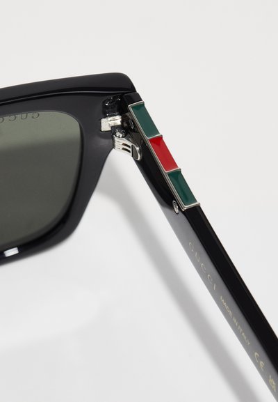 Ochelari de soare din plastic negru cu lentile gri. Temperele au un accent din email verde, alb și roșu. Elemente metalice lângă balama.